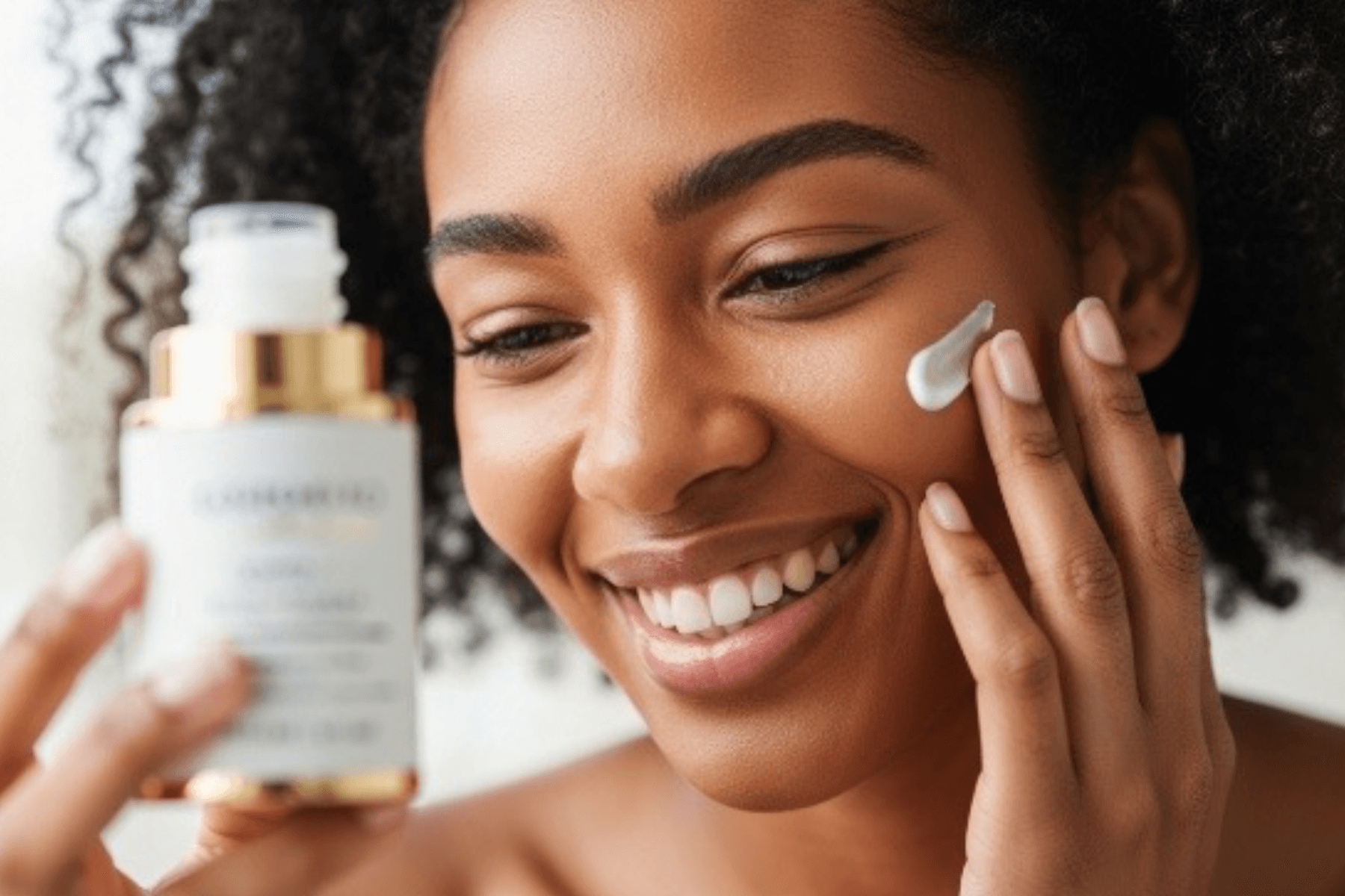 A smiling woman with curly hair applies a cream to her cheek with her right hand, while holding a white and gold pump bottle in her left hand.